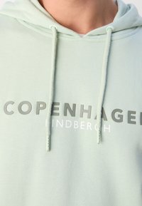 Sweat à capuche vert clair avec cordons, avec inscription en relief "COPENHAGEN" et un plus petit "LINDBERGH" sous le texte principal.
