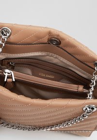 Beige brokattu käsilaukku, jossa on hopeinen ketjuhihna, avoimena näyttää sisäisen vetoketjupussin ja "Steve Madden" -tarran sisällä.