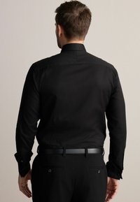Cămașă neagră cu mânecă lungă, cu guler structurat, dintr-un material fin, și cu manșete închise cu nasturi, purtată cu pantaloni închiși la culoare și o curea subțire neagră.