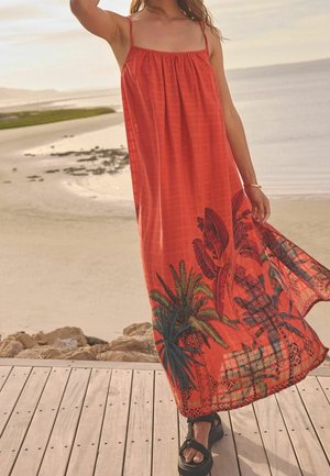 Robe longue rouge à fines bretelles, avec un imprimé tropical de palmes en noir et vert au bas. Tissu léger, design fluide.