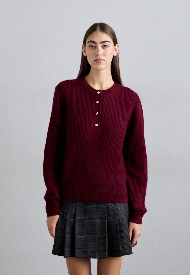 Giovane donna con lunghi capelli castani che indossa un maglione burgundy a coste con bottoni e una gonna plissettata a quadri grigia scura, su uno sfondo semplice.
