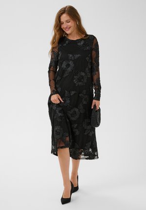 Glimlachende vrouw in een zwarte, lange mouwen bloemen mesh jurk met zwarte hakken houdt een zwarte clutch met structuur vast tegen een effen lichte achtergrond.