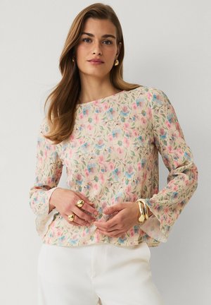 Frau mit langen braunen Haaren trägt eine pastellfarbene, geblümte Langarmbluse, weiße Hose und goldene Ringe und Armbänder, steht vor weißem Hintergrund.