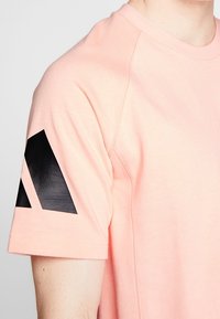 Camiseta rosa claro de manga corta, con un diseño abstracto negro en el hombro izquierdo. Tejido suave con una textura lisa.