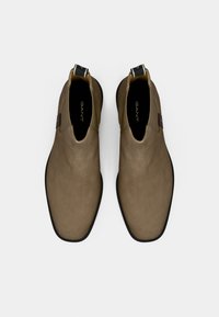 Bottines en suède marron avec une texture lisse et une semelle en caoutchouc noir. Elles possèdent une languette et le logo Gant au niveau du talon. Design classique Chelsea.