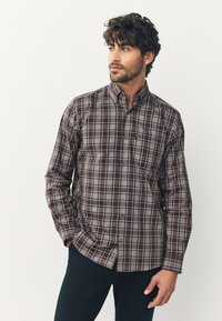 Chemise à carreaux marron à boutons, dotée d'un col, de manches longues, et de lignes contrastantes bleu et blanc, portée avec un pantalon foncé.