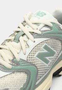 Közeli kép egy fehér és zöld New Balance futócipőről, látható a fűző, a hálós anyag, valamint a logó az oldalon és a nyelvén.