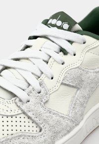 White sneaker with a gray textured suede panel, smooth leather accents, green lace holder, and logo on the tongue. Perforated toe box.
