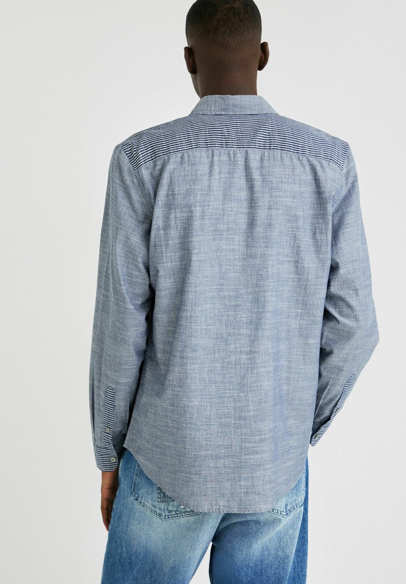 Mann trägt ein langärmliges, hellblaues Hemd mit Muster und gestreiften Details an den Schultern und Manschetten, kombiniert mit hellblauen Jeans.