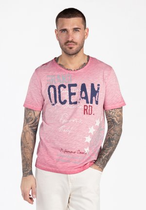 Mann mit kurzen Haaren und tätowierten Armen trägt ein rosa T-Shirt mit der Aufschrift "Grand Ocean Rd." und hellfarbene Hose, steht mit einer Hand in der Tasche.