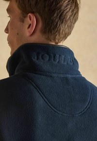 Detailný záber na hornú časť chrbta muža s tmavomodrou flísovou bundou s vysokým golierom, na ktorom je vyrazené slovo "JOULES."