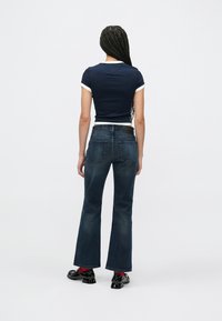 Μπλε κοντομάνικη μπλούζα με λευκή επένδυση, συνδυασμένη με σκούρο τζιν με φαρδιά παντελόνια. Τα παπούτσια του μοντέλου είναι μαύρα γυαλιστερά με τετράγωνη μύτη.