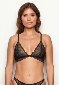 Frau mit langen braunen Haaren trägt einen schwarzen Spitzen-Bralette und passende Spitzenunterwäsche vor einem schlichten weißen Hintergrund.