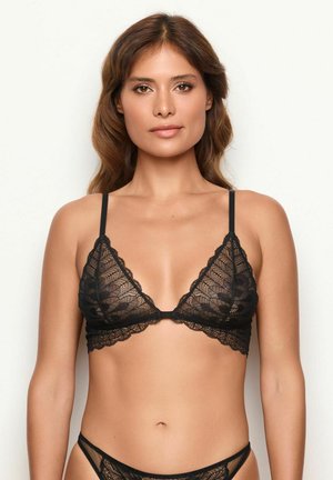 Vrouw met lang bruin haar draagt een zwarte kanten bralette en bijpassend kanten ondergoed tegen een plain witte achtergrond.