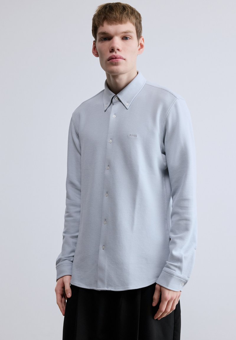 Svetlosivá košeľa s dlhým rukávom s textúrovaným povrchom, golierom na gombíky, prednými gombíkmi a malým logom vyšitým na ľavej strane hrudníka.