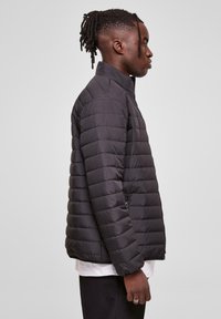 Black padded jacket with a high collar, featuring a zip closure, side pockets, and a quilted texture. Shown from the side.