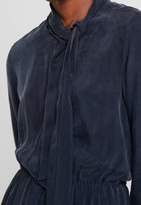 Abito blu navy in tessuto leggero, con colletto annodato, maniche lunghe e vita arricciata per una silhouette morbida.