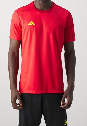 Uomo che indossa una maglia sportiva rossa a maniche corte con logo giallo sul petto e pantaloncini neri con logo giallo abbinato sulla gamba.