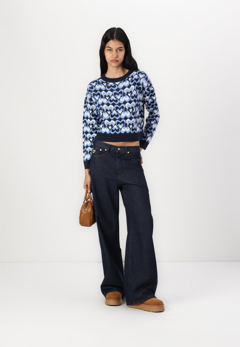 Blauwe gebreide trui met hartpatroon en geribbelde manchetten, gecombineerd met donkere denim wijde broek en bruine platform schoenen. Bruine handtas inbegrepen.