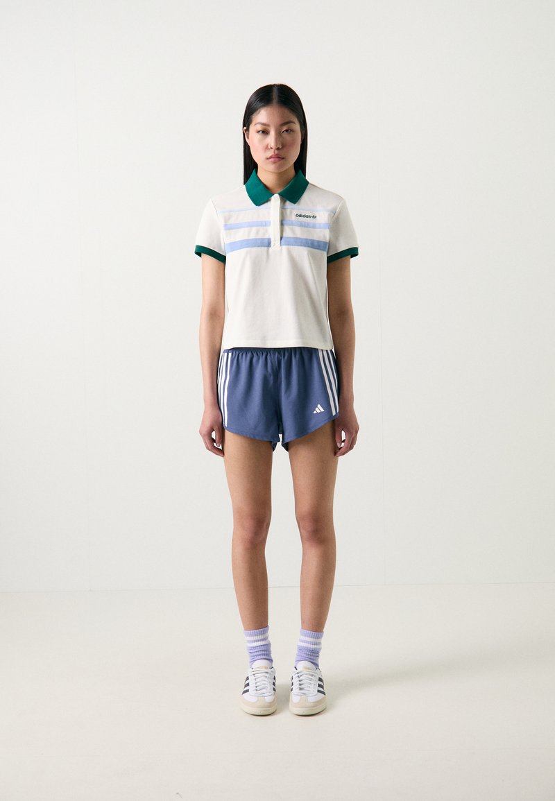 Camisola polo em creme com riscas verdes e azuis, combinada com calções azul-marinho com riscas brancas. Ténis brancos e meias roxas.