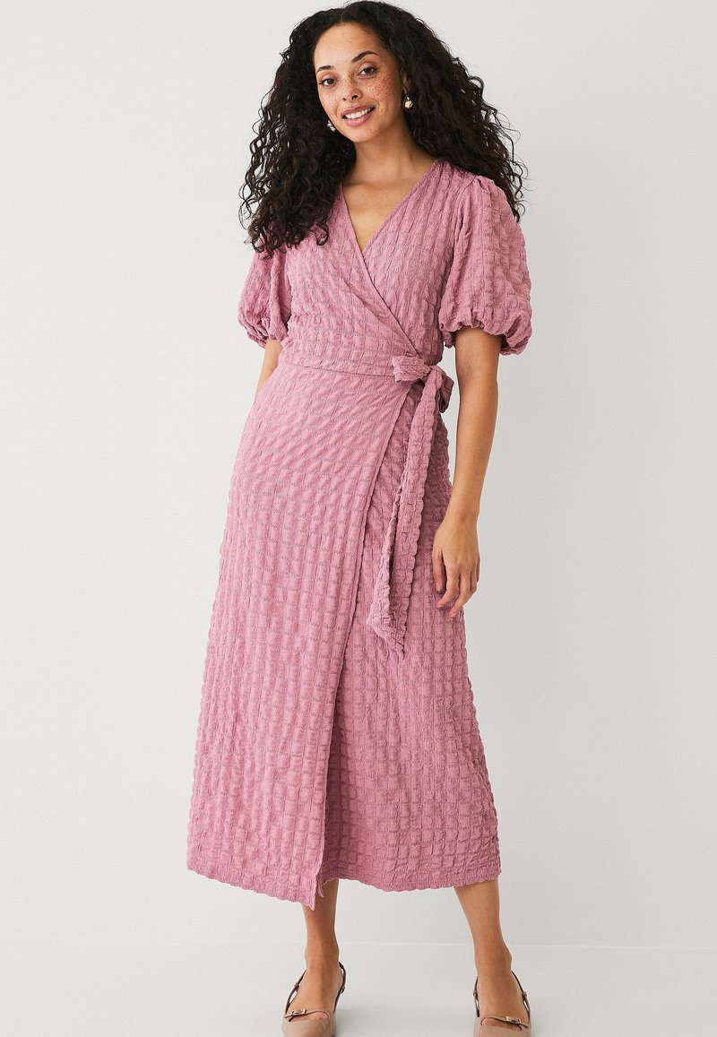 Rosa Wickelkleid mit Puffärmeln, strukturiertem Stoff und einem Bindegürtel in der Taille. Die Länge reicht bis zur Mitte der Wade und hat einen dezenten V-Ausschnitt.