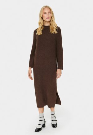 Braunes Midi-Kleid aus Strick mit langen Ärmeln, gerippter Textur, Rundhalsausschnitt, seitlichen Schlitzen, kombiniert mit grauen Socken und schwarzen Blockabsatzschuhen.