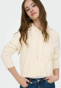 Jeune femme aux longs cheveux bruns portant un sweat à capuche crème avec fermeture éclair et un jean bleu, posant les mains dans les poches devant un fond uni.