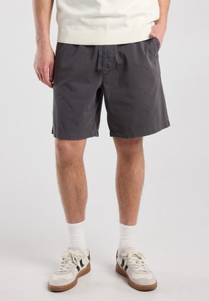 Man wearing dark gray shorts, white socks, and white sneakers with black details, standing with one hand in pocket.