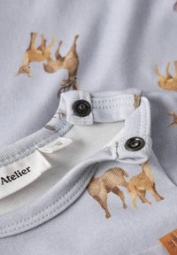 Camicia in cotone azzurro chiaro con animali marroni illustrati. Chiusure a scatto sul collo. Texture morbida e bordi a costine. Etichetta visibile.