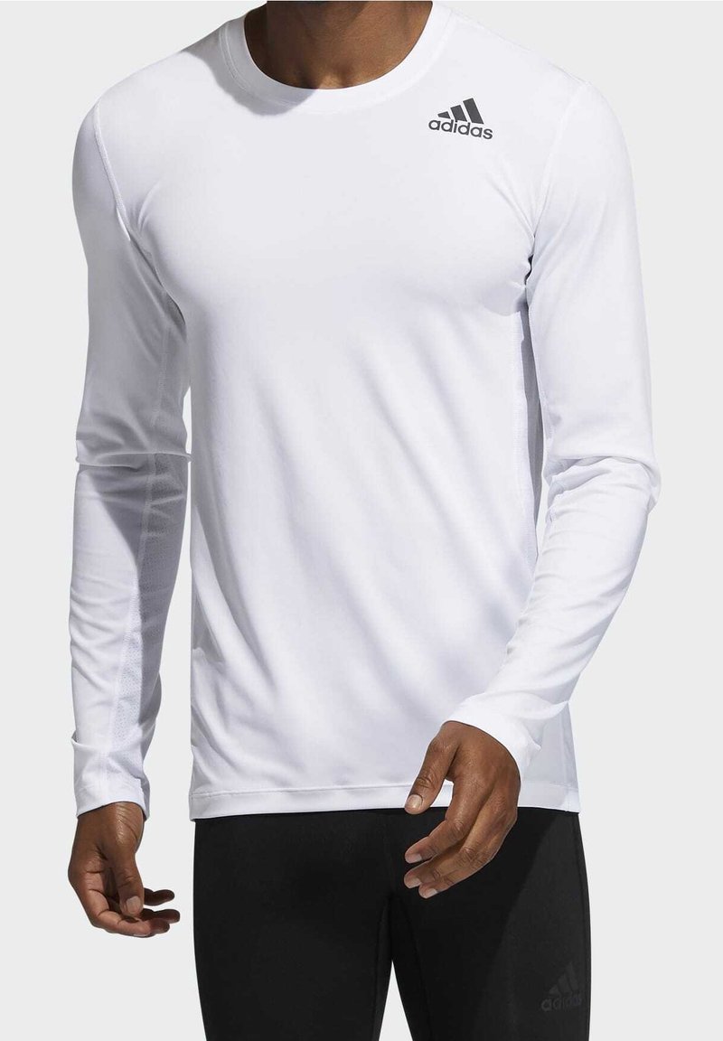 Långärmad vit träningsskjorta gjord av slät tyg med rund hals; har en liten svart Adidas-logotyp på övre vänstra sidan.