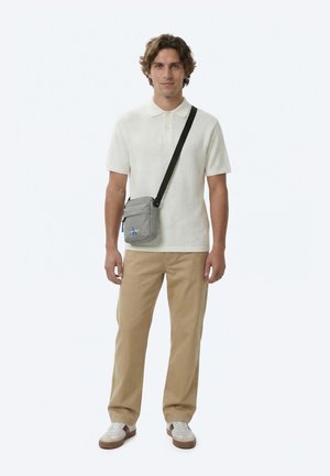 Muž oblečený v bielej pletenej polo košeli s krátkym rukávom, béžových nohaviciach, bielych teniskách a so sivou taškou cez rameno s čiernym popruhom stojaci na bielom pozadí.