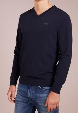 Pull à col V marine en tissu tricot doux, doté de manches longues et d'une coupe ajustée. Un logo discret sur la poitrine ajoute une touche de détail.