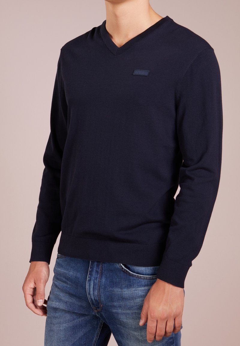 Maglione a V in blu navy in morbido tessuto a maglia, con maniche lunghe e silhouette aderente. Un logo discreto sul petto aggiunge un tocco di dettaglio.