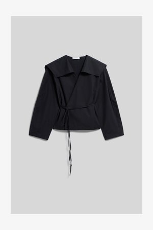 Blouse noire à manches longues, col large et détail de nouage sur le côté, posée à plat sur un fond gris clair.