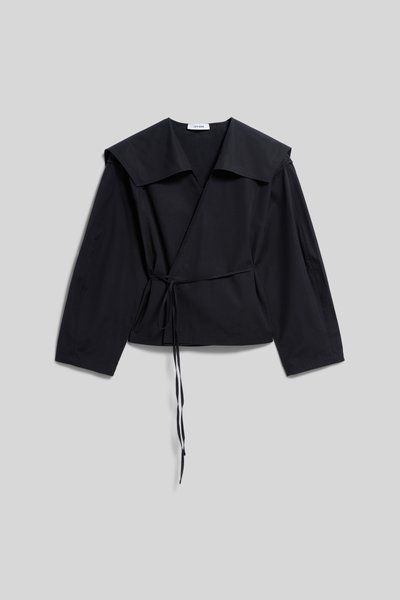 Blouse noire à manches longues, col large et détail de nouage sur le côté, posée à plat sur un fond gris clair.