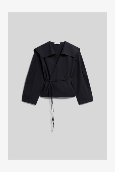 Blouse noire à manches longues, col large et détail de nouage sur le côté, posée à plat sur un fond gris clair.