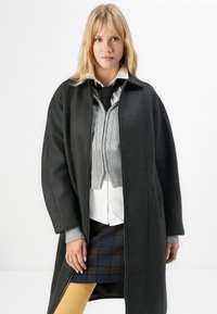 Blondinka v črnem plašču, ki ga odpre, da pokaže sivo pleteno brezrokavnik, belo srajco, črno kravato, karirano krilo in rumene nogavice, stoji pred belo ozadjem.