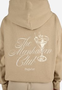 Bežinė kapučinų striukė su švelnia tekstūra, su balta užrašu "The Manhattan Club" ir iliustracija kokteilių stiklo.