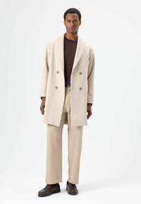 Beige wollen jas met dubbelgeknoopt ontwerp, grote knopen, gedragen over een donkerbruin overhemd en beige broek, aangevuld met donkerbruine schoenen.