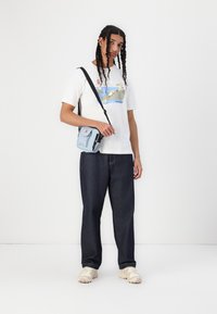 T-shirt branco com gráfico colorido, calças de ganga escuras, mala de ombro azul clara e sapatilhas bege. Modelo tem cabelo longo e trançado.