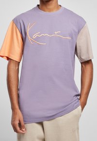 Fialové tričko s krátkým rukávem s oranžovými a béžovými akcenty; na přední straně se nachází vyšitý název značky v kontrastní barvě.