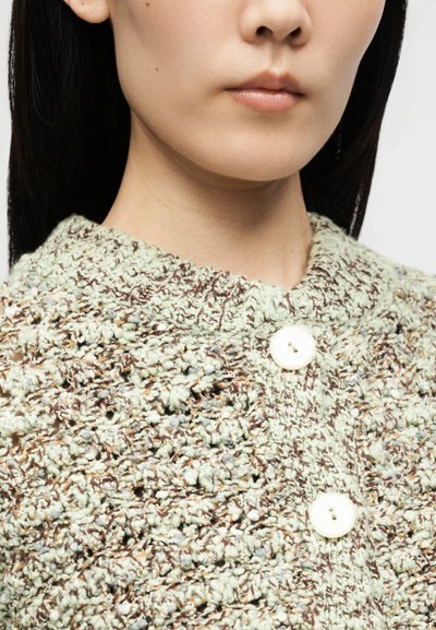 Gros plan d’une personne portant un cardigan en maille texturée avec de gros boutons blancs et un col côtelé, montrant le bas du visage et le cou.