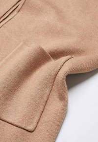 Tissu en tricot beige montrant une texture douce avec un petit détail de poche ; le matériau semble léger et chaud, présentant un motif côtelé subtil.