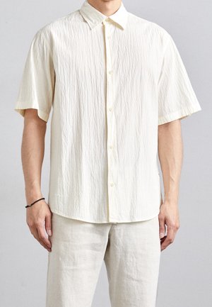 Homme portant une chemise à manches courtes texturée de couleur blanc cassé à boutons et un pantalon beige clair, devant un fond uni.