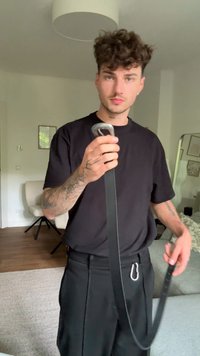 Cinto de couro preto com uma fivela de metal prata, segurado por uma pessoa com tatuagens. O fundo apresenta uma cadeira de cor neutra e decoração.