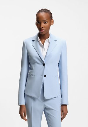 Ung kvinde i lyseblåt skræddersyet jakkesæt og hvid skjorte, stående mod en ensartet hvid baggrund, vendt fremad med et neutralt udtryk.