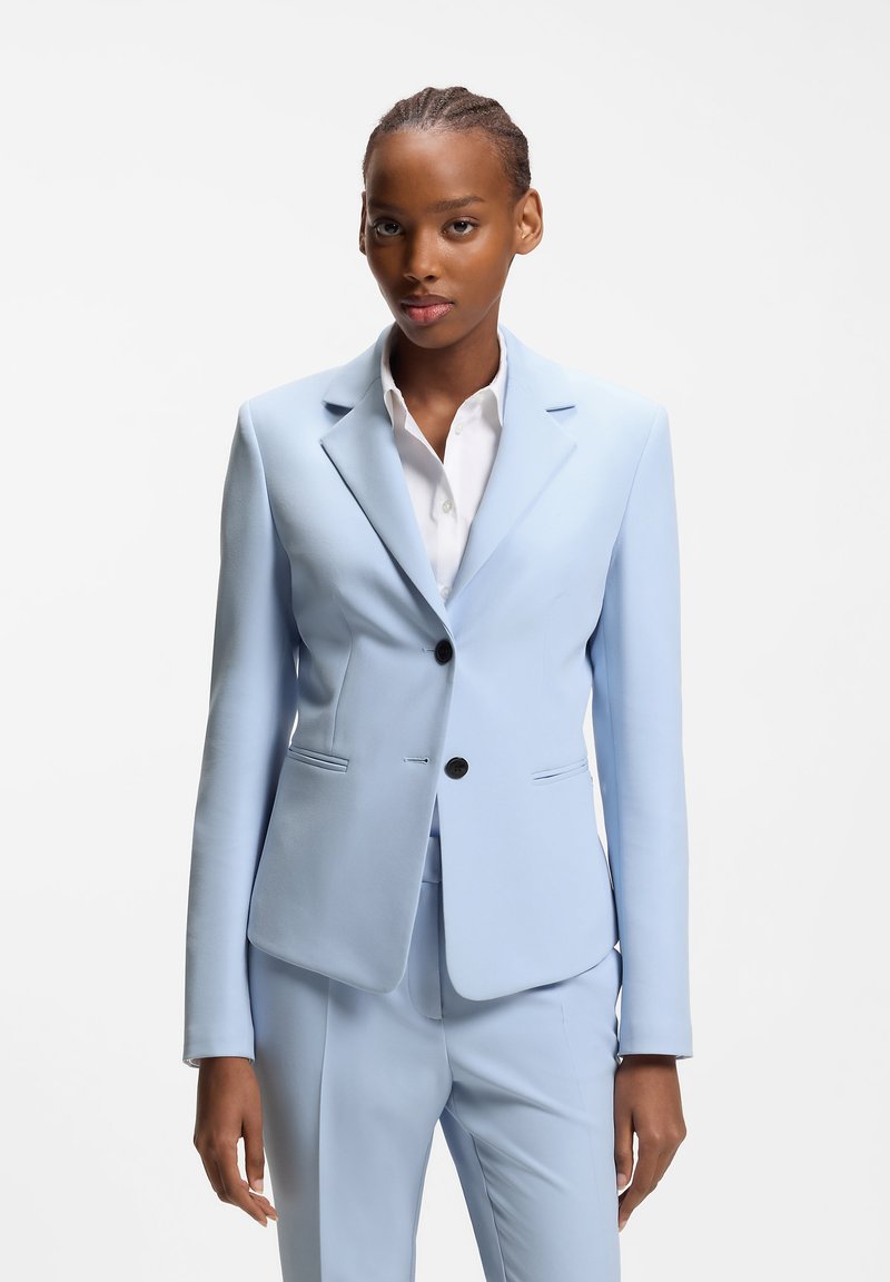 Ung kvinde i lyseblåt skræddersyet jakkesæt og hvid skjorte, stående mod en ensartet hvid baggrund, vendt fremad med et neutralt udtryk.