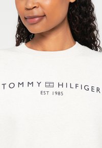 Lysfarget hettegenser med "TOMMY HILFIGER" i fet marineblå tekst; rund hals og myk, teksturert stoff med etablert dato.