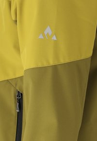 Keltainen ja oliivinvihreä takin hiha, jossa on musta vetoketju ja valkoinen kolmionmuotoinen logo yläosassa.