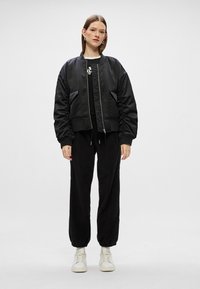 Czarna kurtka bomberka z zapięciem na zamek, ściągaczami przy mankietach i bocznymi kieszeniami, w połączeniu z czarnymi dresami i białymi trampkami. Gładka, matowa tekstura.